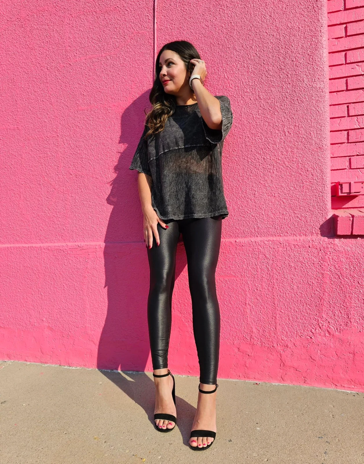 Black faux-leather leggings and a dark blouse against a pink brick wall, heeled sandals, sunlight from the left