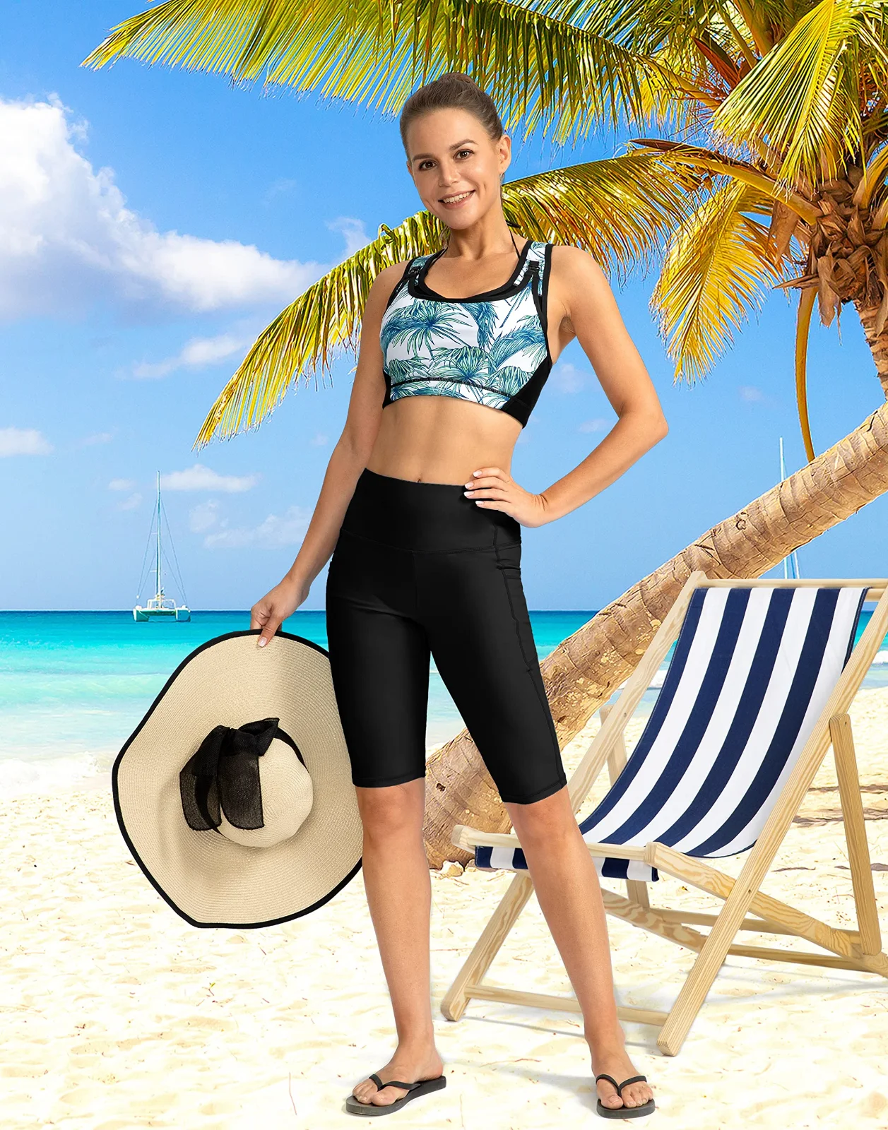 Black knee-length swim shorts and a patterned swim top on a beach under palms, a straw hat in one hand